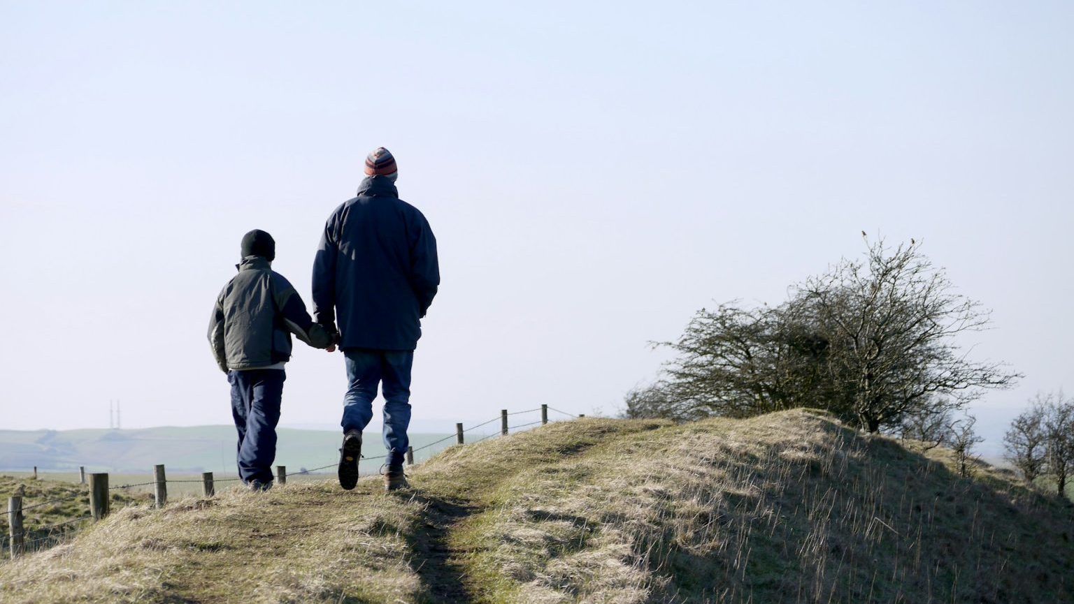 Two people walk hand in hand on a grassy hilltop trail, wearing winter jackets and hats. Sparse trees and a distant landscape are visible under a clear sky.