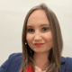 A person with long straight hair, wearing a navy blazer and red shirt, looks at the camera against a plain background. They have a small microphone attached near their left ear.